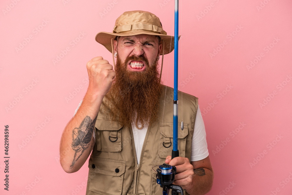 Young caucasian ginger fisherman with long beard holding a rod isolated on pink background showing fist to camera, aggressive facial expression. Young caucasian ginger fisherman with long beard holding a rod isolated on pink background showing fist to camera, aggressive facial expression.