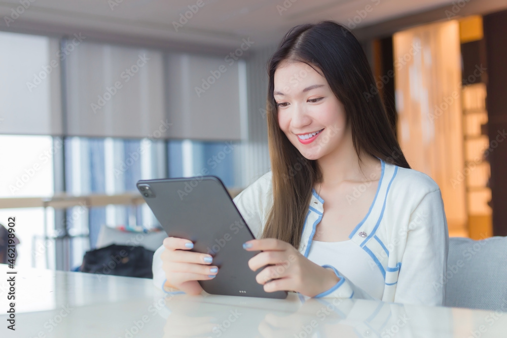 Beautiful adult young age Asian professional woman with long hair in white shirt holds tablet her smiling happily while she sits on chair in workplace office with glassed building as a background. Beautiful adult young age Asian professional woman with long hair in white shirt holds tablet her smiling happily while she sits on chair in workplace office with glassed building as a background.