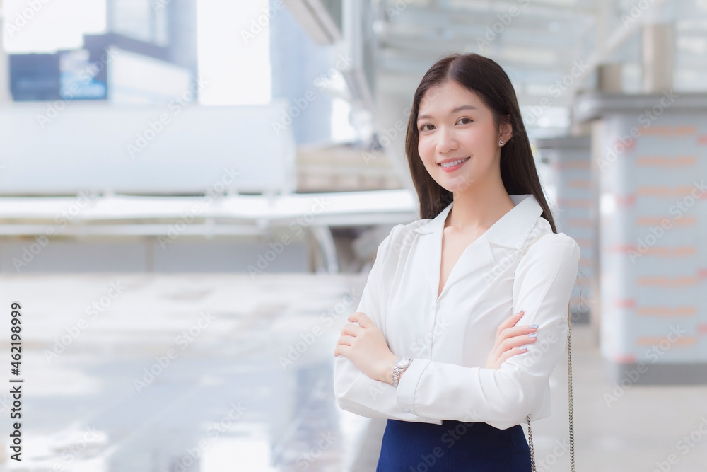 Asian confident working woman who has a long hair with a white shirt is standing and arm crossing  standing urban outdoors while walking to office in big city with business buildings.