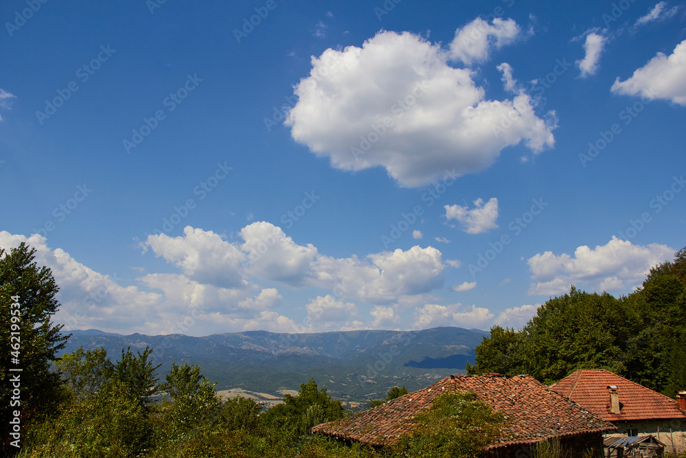 Fluffy clouds in an azure sky over forest, mountains, green vegetation. Tiled roof of an old country house.