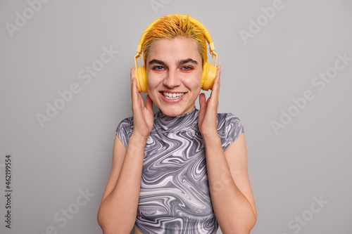 Happy hipster girl with trendy yellow hairstyle bright makeup listens music in wireless headphones has cheerful mood enjoys favorite rock song dressed casually isolated over grey background.
