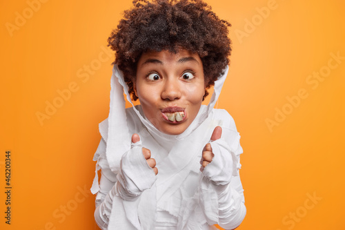 Isolated shot of funny curly haired young African American woman has funny grimace shows false teeth keeps thums up dresses for halloween party isolated over orange background. 31st October is coming photo