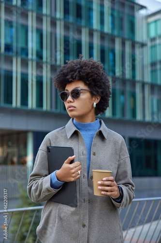 Stylish Afro American woman walks on city street with digital tablet and paper cup of coffee uses modern technologies wears sunglasses and jacket poses against modern buildings. People and lifestyle