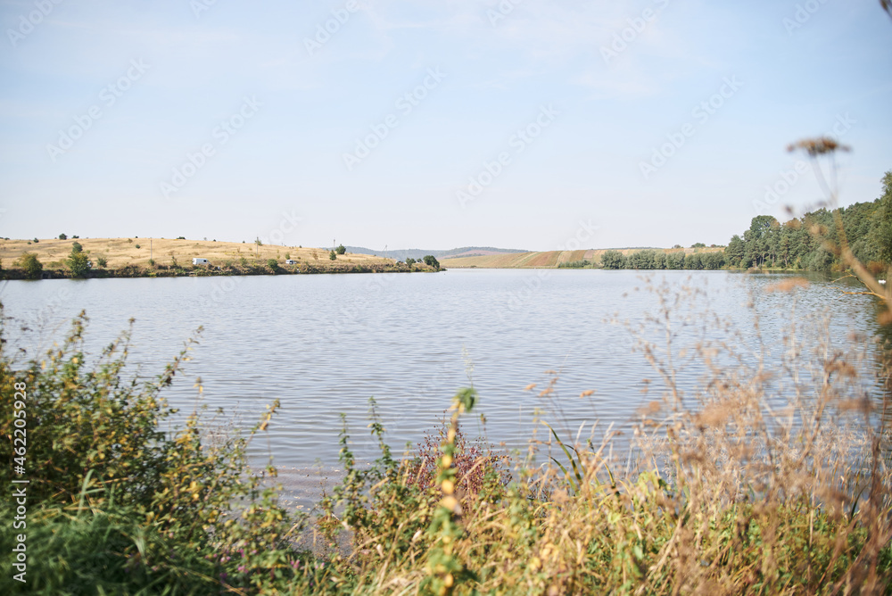 Amazing lake landscape with dry grass and green trees in autumn.Light blue sky over pond. Nature protection concept. Rural scene in countryside. Fish farming basin in the village. Amazing lake landscape with dry grass and green trees in autumn.Light blue sky over pond. Nature protection concept. Rural scene in countryside. Fish farming basin in the village.