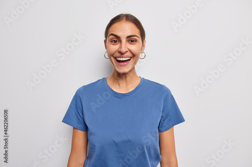 Wallpaper Mural Studio shot of happy dark haired young woman smiles broadly expresses positive emotions dressed in casual blue t shirt isolated over white background. Cheerful female model reacts on funny news Torontodigital.ca