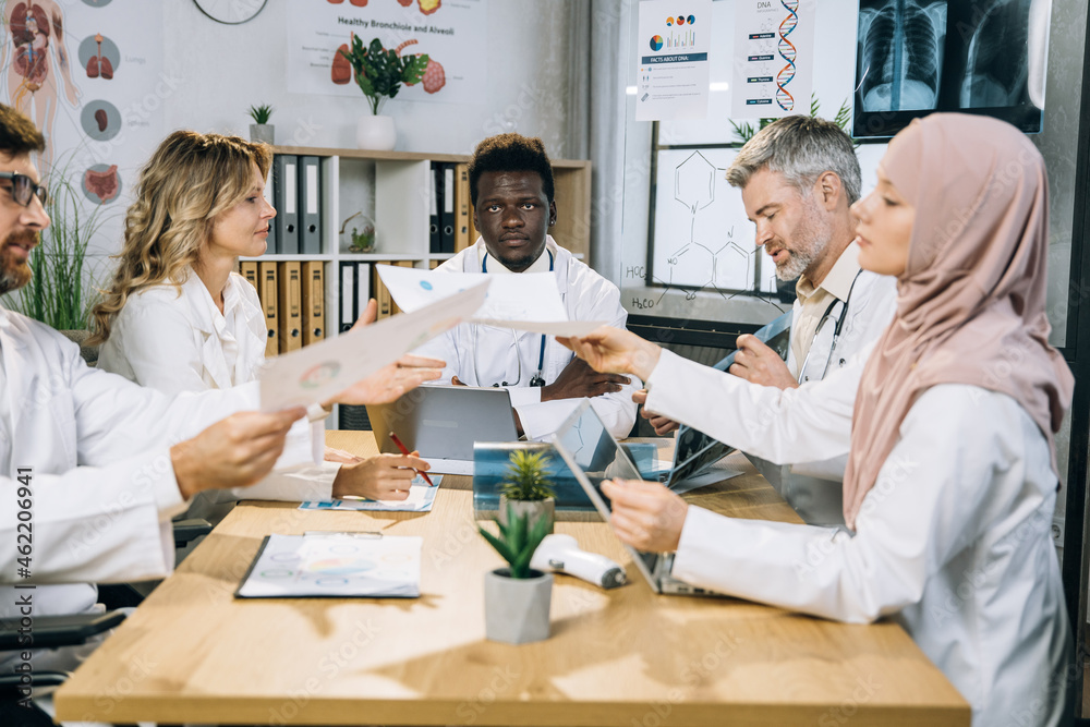 Team of mixed race doctors sitting at table on conference room and sharing with reports during medical research. Male and female therapists cooperating for common success. Team of mixed race doctors sitting at table on conference room and sharing with reports during medical research. Male and female therapists cooperating for common success.
