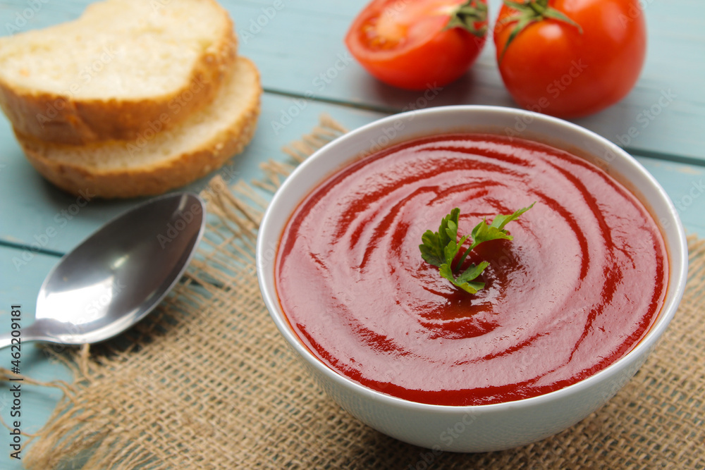 Tomato soup with herbs with bread in a white bowl with a spoon on a blue wooden table. Tasty food. Tomato soup with herbs with bread in a white bowl with a spoon on a blue wooden table. Tasty food.