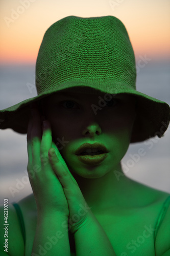 Trendy curvy model with a hat in green neon light with a sunset in background. Eyes not visible.