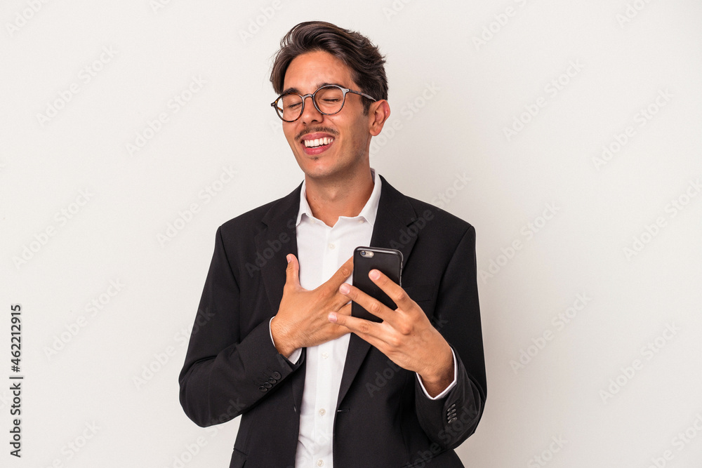 Young mixed race business man holding mobile phone isolated on white background laughs out loudly keeping hand on chest. Young mixed race business man holding mobile phone isolated on white background laughs out loudly keeping hand on chest.