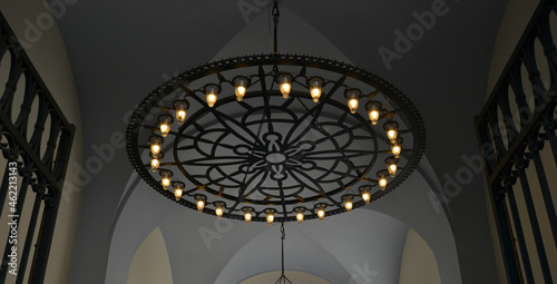 Round ornamental chandelier in black with light bulbs hanging from ceiling behind open gates.