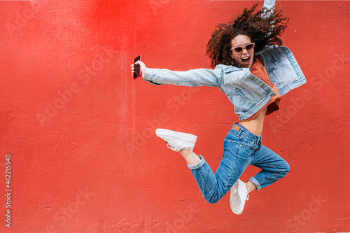 Excited young woman wearing sunglasses jumping in front of red wall