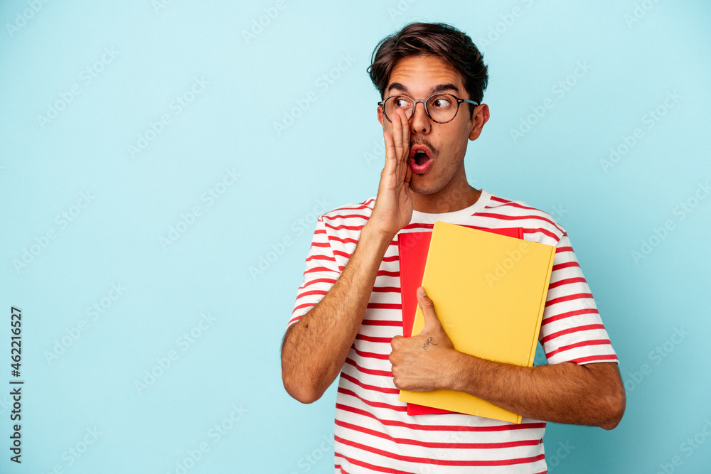 Young mixed race student man holding books isolated on blue background is saying a secret hot braking news and looking aside Young mixed race student man holding books isolated on blue background is saying a secret hot braking news and looking aside