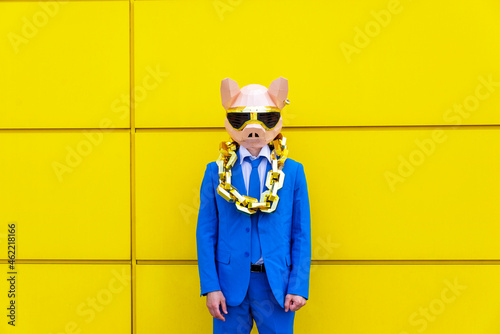 Man wearing vibrant blue suit, pig mask and large golden chain standing in front of yellow wall photo