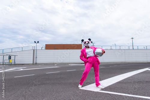 Woman wearing vibrant pink suit and panda mask posing in empty parking lot with disco ball in hand photo