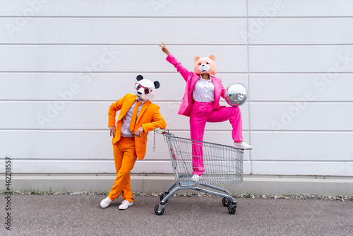 Man and woman wearing vibrant suits and animal masks posing with shopping cart and disco ball in front of white wall photo