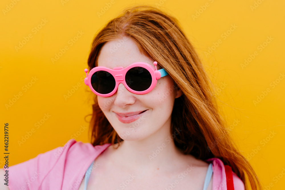 Woman wearing pink sunglasses smiling in front of yellow wall Woman wearing pink sunglasses smiling in front of yellow wall