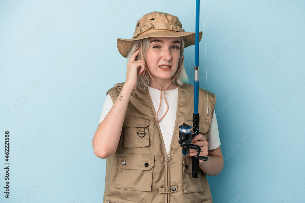 Young caucasian fisherwoman holding a rod isolated on blue background covering ears with hands. Young caucasian fisherwoman holding a rod isolated on blue background covering ears with hands.