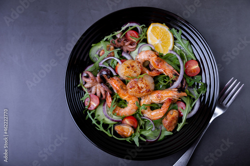 Salad with seafood, arugula, tomatoes, cucumbers, red onion and lemon on a black plate. Black background, close-up.