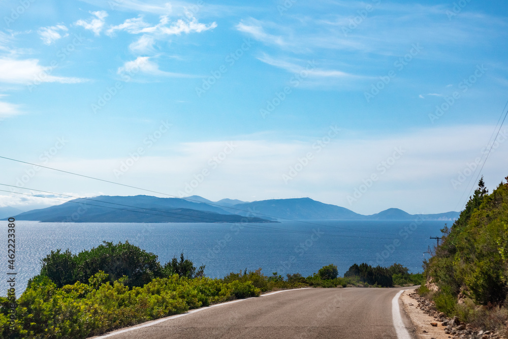 Obraz premium Driving high steep road with green vivid hills, blue distant islands silhouettes and clear blue sky. Travel Lefkada island, Greece. Sunny summer scenic day trip