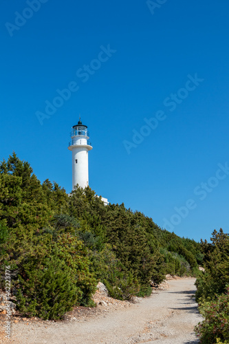 Ionian sea island lighthouse on green cliff with dirt road on a bright clear blue day in Greece. Scenic travel destination. Lefkada island. Vertical