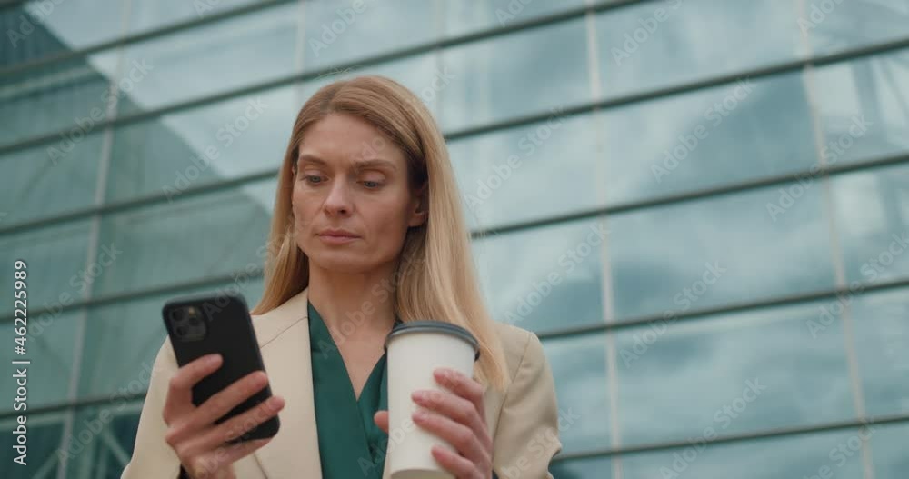 Serious Successful Middle-Aged Woman Office Manager looking at Phone Screen. Standing near Glass Business Center holding a cup of coffee. Handsome Caucasian Businesswoman uses Mobile Apps for Chatting Serious Successful Middle-Aged Woman Office Manager looking at Phone Screen. Standing near Glass Business Center holding a cup of coffee. Handsome Caucasian Businesswoman uses Mobile Apps for Chatting