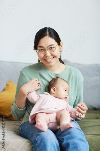 Portrait of Asian young woman in eyeglasses smiling at camera while sitting on the bed with her little daughter on her knees