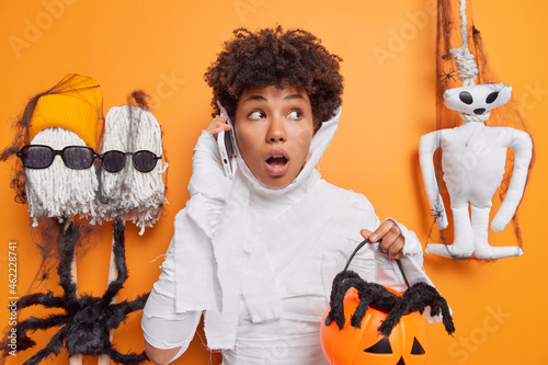 Shocked ethnic woman dressed like ghost for halloween party holds carved pumpkin makes telephone call looks away with scared expression celebrates mystery event invites friends for celebration