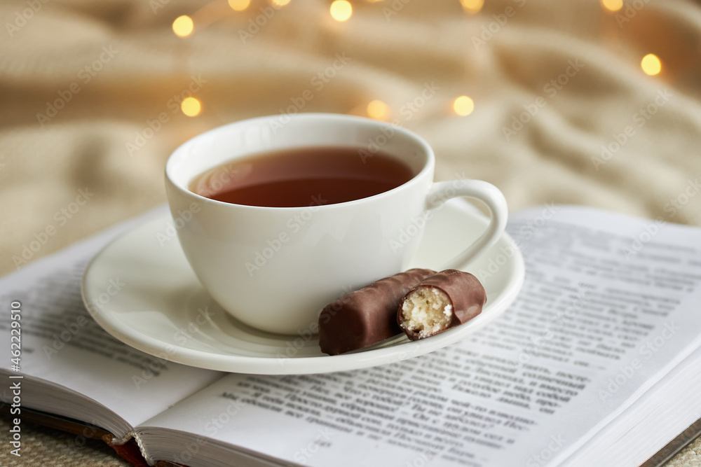 White cup of tea with chocolate sweets on a n open book and beige plaid in the Christmas lights. White cup of tea with chocolate sweets on a n open book and beige plaid in the Christmas lights.