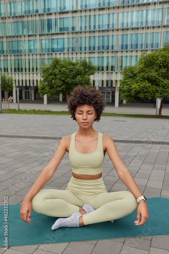 Relaxed sporty young woman sits crosses legs on mat practices yoga enjoys meditation keeps eyes closed breathes deeply poses on fitness mat against urban setting. Healthy habit and axiety relief