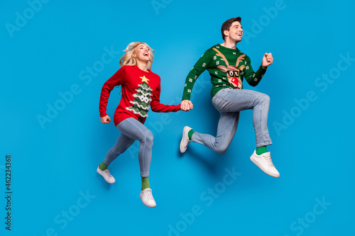 Photo of cheerful pretty young couple dressed ugly print sweaters jumping high holding arms empty space isolated blue color background