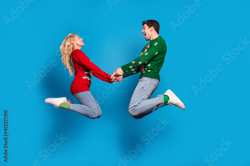 Photo of impressed sweet young people wear ugly ornament pullovers jumping high holding arms isolated blue color background