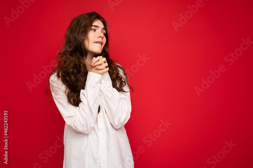 Portrait of beautiful young curly brunette woman wearing white shirt isolated on red background with free space, wanting and dreaming with sincere emotions