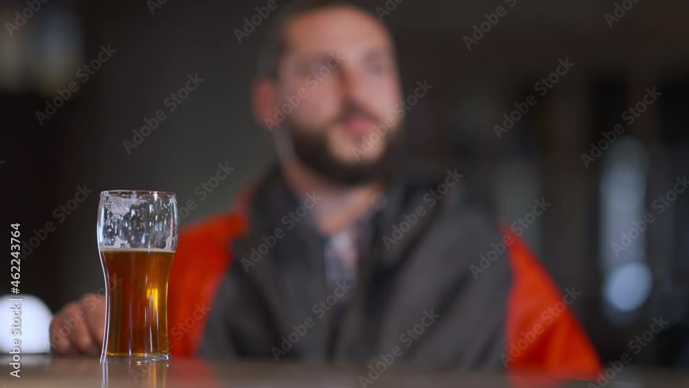 Beer glass on bar counter with blurred man watching sport match rejoicing success shaking German flag. Positive excited Caucasian bearded brutal man making victory gesture in pub Beer glass on bar counter with blurred man watching sport match rejoicing success shaking German flag. Positive excited Caucasian bearded brutal man making victory gesture in pub