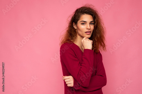 Photo of young european thoughtful charming cute brunette curly woman with sincere emotions wearing trendy pink sweater isolated over pink background with free space