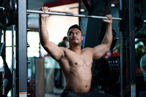 Shirtless adult Asian men sweating while lift up the barbell workout machine for muscle part inside of fitness gym. Bodybuilding athlete sport training for body strength and good health.