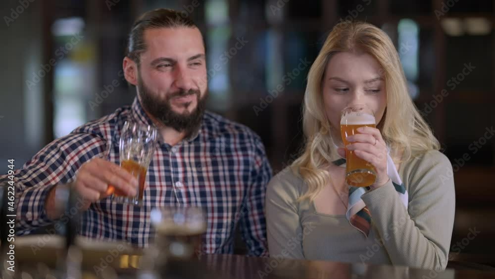 Front view positive man and woman toasting looking at camera drinking beer sitting at bar counter. Portrait of Caucasian friends or couple enjoying weekend leisure posing in pub indoors with alcohol Front view positive man and woman toasting looking at camera drinking beer sitting at bar counter. Portrait of Caucasian friends or couple enjoying weekend leisure posing in pub indoors with alcohol