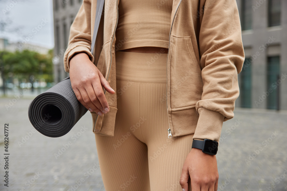Cropped shot of unrecognizable athletic woman in sportsclothes and fitness watch carried rolled up karemat going to have yoga practice and exercise outside poses outside against blurred background