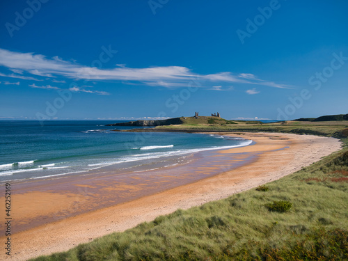 A view of the pristine sands and blue water of the  Northumberland coast in England, UK. Taken on a sunny day with blue skies and white clouds in  summer. Dunstanburgh Castle appears in the distance. © Alan