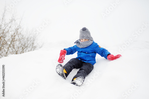 Little boy enjoy riding on ice slide on snowy day. Baby having fun during blizzard. Outdoor winter activities for kids.