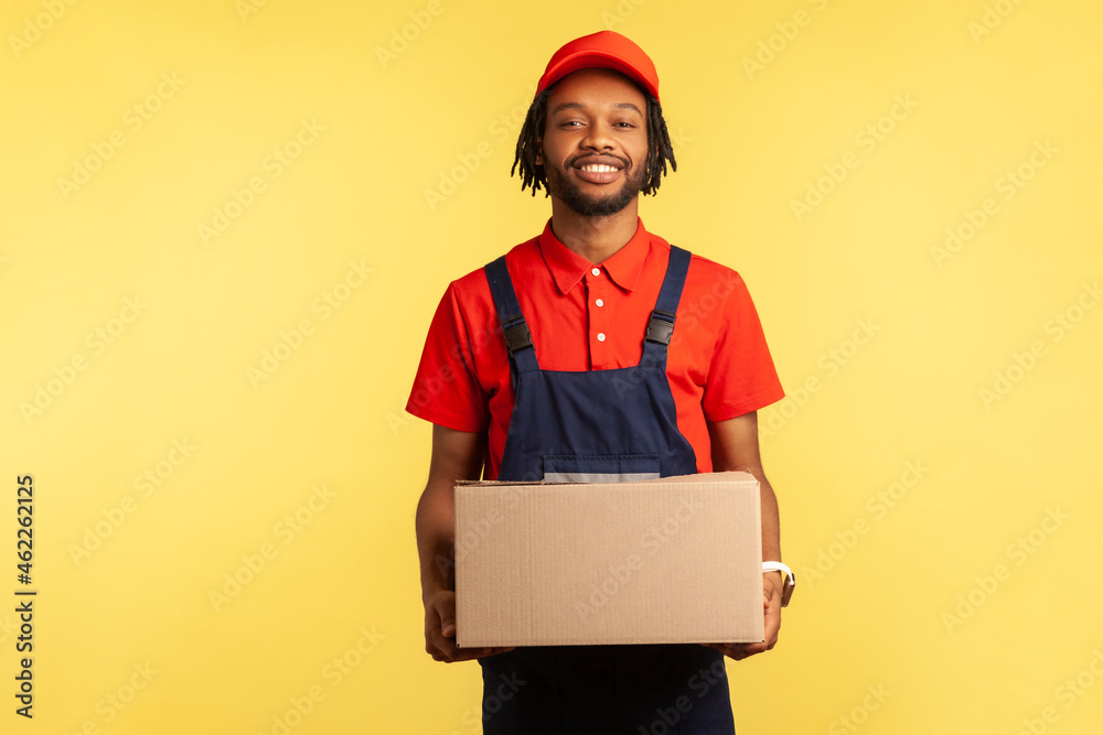 Smiling positive courier wearing blue overalls holding cardboard parcel, delivering orders to address, looking at camera with friendly expression. Indoor studio shot isolated on yellow background. Smiling positive courier wearing blue overalls holding cardboard parcel, delivering orders to address, looking at camera with friendly expression. Indoor studio shot isolated on yellow background.
