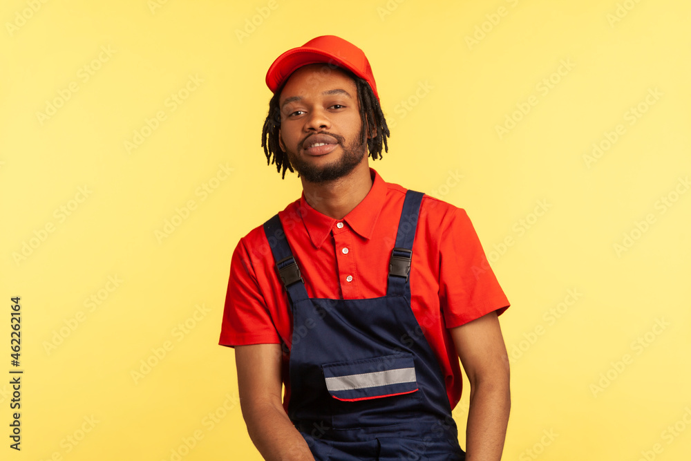 Portrait of bearded handyman with pleasant appearance wearing blue overalls, res T-shirt and visor cap, posing looking directly at camera. Indoor studio shot isolated on yellow background. Portrait of bearded handyman with pleasant appearance wearing blue overalls, res T-shirt and visor cap, posing looking directly at camera. Indoor studio shot isolated on yellow background.