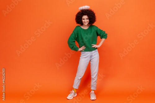 Full length of confident angelic woman with Afro hairstyle wearing green casual style sweater standing with hands on hips, having nimb over her head. Indoor studio shot isolated on orange background.