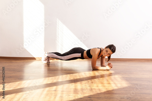 Happy positive athletic female doing plank exercise, looking away with smile, enjoying sport, wearing black sports top and tights. Full length studio shot illuminated by sunlight from window.