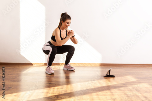 Athletic female doing squats while looking online fitness class, learning correct exercise technique, wearing black sports top and tights. Full length studio shot illuminated by sunlight from window.