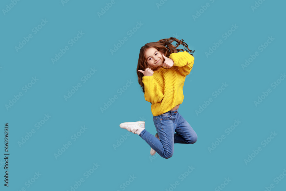 Full length portrait of happy charming little girl jumping up high and showing thumb up to camera, wearing yellow casual style sweater. Indoor studio shot isolated on blue background. Full length portrait of happy charming little girl jumping up high and showing thumb up to camera, wearing yellow casual style sweater. Indoor studio shot isolated on blue background.