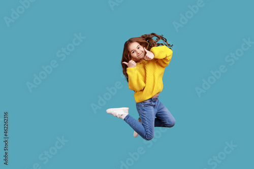 Fotografia Full length portrait of happy charming little girl jumping up high and showing thumb up to camera, wearing yellow casual style sweater