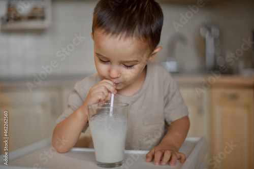 A three-year-old little boy drinks milk from a glass in the kitchen through a paper tube