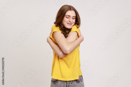 I am egoist, love myself! Selfish narcissistic teen girl with wavy hair embracing herself and smiling, closed eyes with dreamy satisfied expression. Indoor studio shot isolated on gray background.