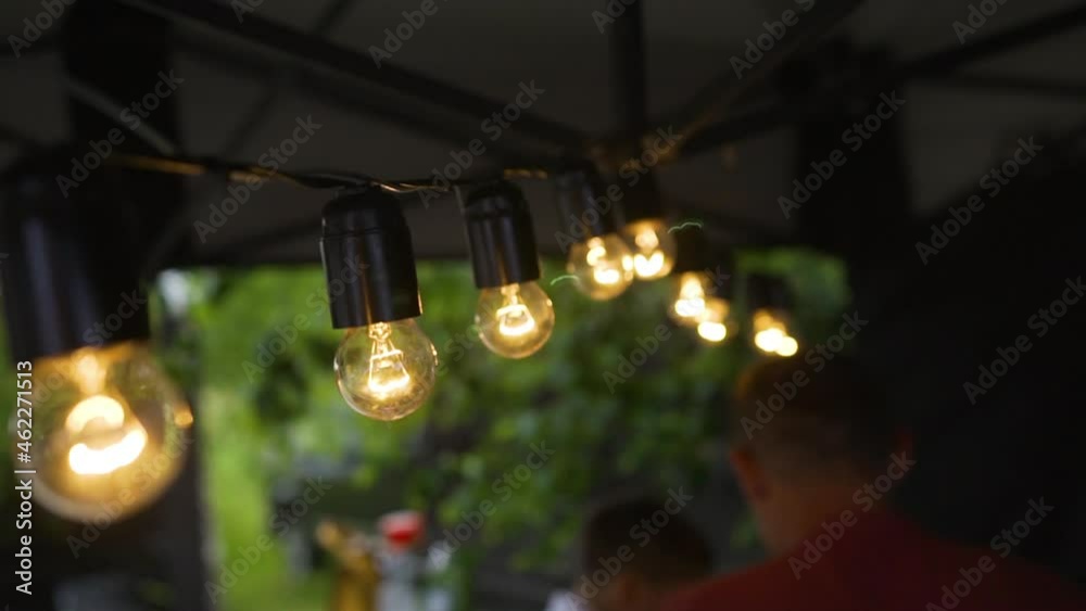 The cord with the light bulbs on hangs in the tent and illuminate it in the evening and as a decorative element. Light bulbs sway in the wind. People walk in the background.