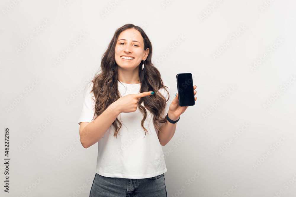 Friendly young woman is pointing at the phone she is holding.